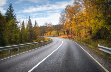Fototapeta premium Road in autumn forest. Beautiful empty mountain roadway, trees with orange foliage and overcast sky. Landscape with asphalt road through the woods in fall. Travel in europe. Road trip. Transportation