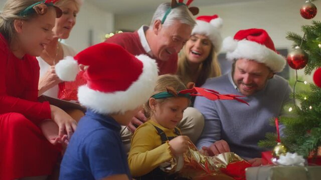Girl unwrapping present of socks as multi-generation family exchange gifts around christmas tree at home - shot in slow motion