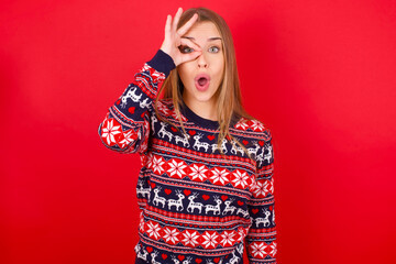 Young caucasian girl wearing christmas sweaters on red background doing ok gesture shocked with surprised face, eye looking through fingers. Unbelieving expression.