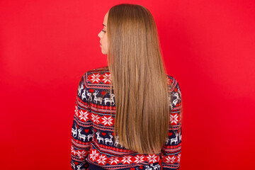 The back side view of a Young caucasian girl wearing christmas sweaters on red background. Studio...