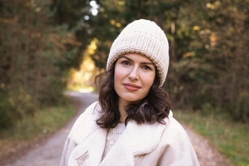 Portrait of a young brunette woman in a knitted hat and white jacket in the autumn forest