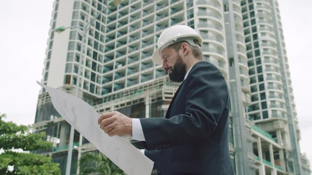 Engineer in white protective helmet checks a construction plan in new microdistrict of the city.Senior Engineer Planning Manufacture Work business suit a red jacket with construction card in his hands