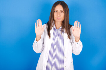 Serious Young european doctor woman on blue background pulls palms towards camera, makes stop...