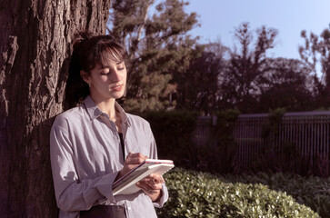 Fototapeta premium Young woman writes in a notebook leaning against the trunk of a tree, and the afternoon sun illuminates her.