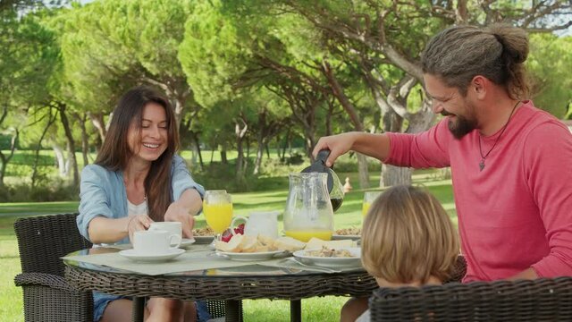 Lovely parents and a kid enjoy their summer vacation at luxury hotel terrace. Cheerful family having tasty breakfast within picturesque natural landscape outdoors. High quality 4k footage