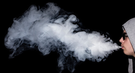 Man iin a hood and sunglasses vaping e-cigarette and blowing a cloud on black background.