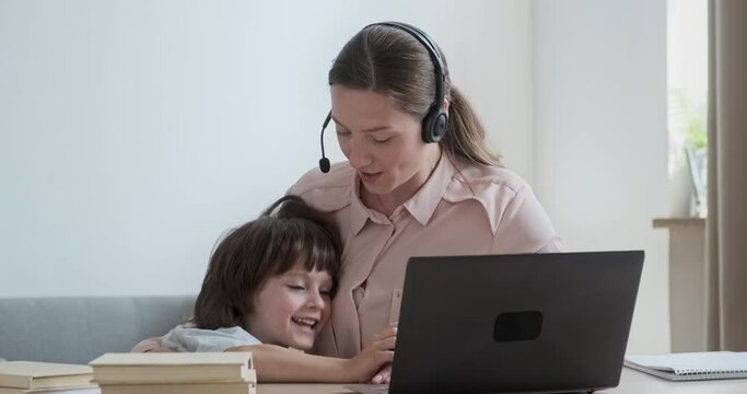 Mother businesswoman working remotely from home on video conference when cute boy rushes up and distracts and upsets mother. Difficulties of working and studying online with small children.