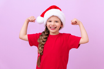 A beautiful little girl in a red Christmas hat and a long pigtail shows her muscles and smiles.