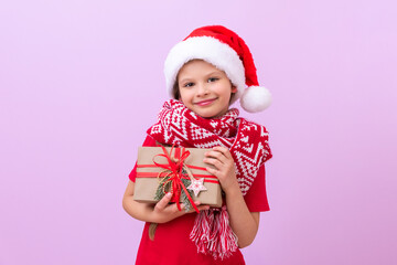 A little girl in a red Christmas scarf and a festive hat holds her Christmas gift and smiles.