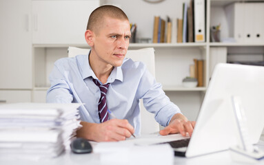 Portrait of manager dressed in the shirt with a tie working with documents and laptop at the modern office interior