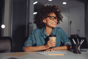 Smiling young multiethnic lady sitting at work desk and holding cup of coffee in the office