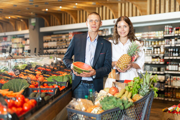 Portrait of a happy couple with a grocery cart in a supermarket, holding a watermelon and pineapple in their hands