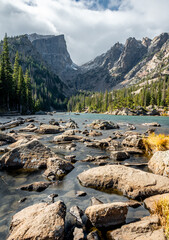 View of dream lake surrounded by mountains in rocky mountain national park, colorado. The lake has many rocks and the peak of the mountain is pointed and surrounded by trees.