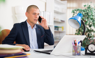 Focused office employee sitting at workplace with laptop and talking on mobile phone