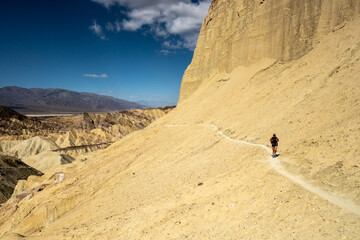 Woman Walks Along Thin Trail Cutting Through Slopes of Death Valley Badlands