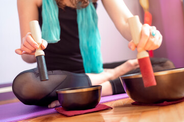 Low section of a young caucasian woman playing tibetan singing bowls over a purple yoga mat on wooden floor. Concept of meditation and relaxation at home. Yogini. Selective focus - Powered by Adobe