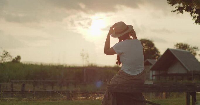 A female tourist in a brown hat, white T-shirt and brown skirt standing on the meadow use her mobile phone to take pictures of the sunset, beautiful orange flare on the farmer village at countryside. 