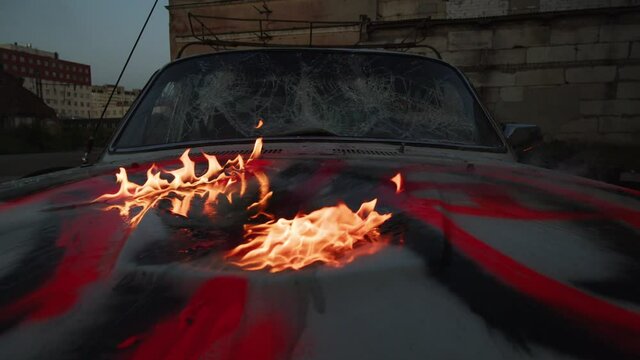 Close look on maliciously wrecked during violent streets demonstrations, car's broken and destroyed details from an angle of bright pressure paint riot signed bonnet covered by blazing fire 