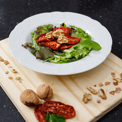 Close-up of plate with salad of lettuce, arugula, sundries tomatoes and walnuts. On wooden board decorated with whole walnuts and sundries tomatoes. Vegetarian or vegan food. Healthy eating concept.