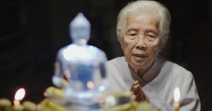Asian Buddhist old woman with white hair white dress praying and raise their hands in front of the glass Buddha image and light candles in the night.