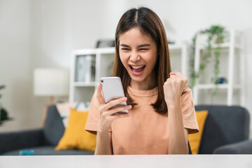 Cheerful beautiful Asian woman holding smartphone with fists clenched celebrating victory expressing success.