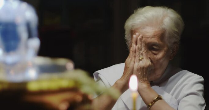 Asian Buddhist old woman with white hair white dress praying and raise their hands in front of the glass Buddha image and light candles in the night.
