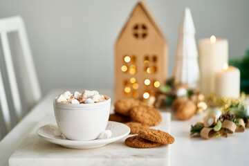 Cozy evening wintertime scene. Christmas holiday background with fir branch, cup of hot cocoa and marshmallow, candles on table. Kraft cozy paper house. Winter hot drink, selective focus