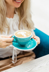 Young woman holding blue cup with cappuccino in her hands and smiling