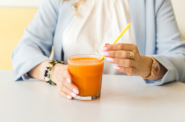A glass of freshly squeezed carrot juice in the hands of a young woman in a blue jacket and white blouse