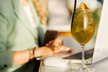 A transparent glass with apple cider stands on the bar counter of a summer cafe next to a laptop, on which a woman in a jacket works