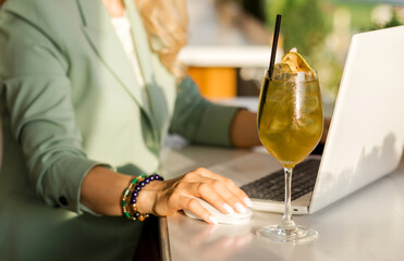 A transparent glass with apple cider stands on the bar counter of a summer cafe next to a laptop,...