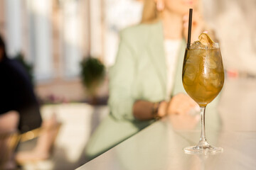 A transparent glass with apple cider stands on the bar counter of a summer cafe. In the background sits a woman with a laptop