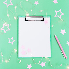 Christmas holidays concept, preparation for xmas, mock up white sheet for wish list. Flat lay style with New Year decoration, glowing garland, pink stars and notebook.
