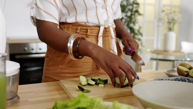 Midsection slowmo shot of unrecognizable hands of African-American woman cutting fresh cucumbers on wooden cutting board at kitchen
