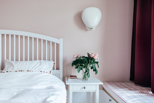 White bed next to pink roses in a vase over a table