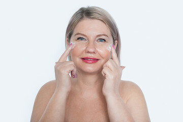 Happy 40s mid aged mature blonde lady applying facial cream on face looking at camera isolated on white background. Anti age healthy dry skin care beauty