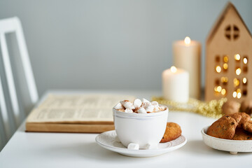 Reading book on cozy winter evening with candles, cocoa hot chocolate with marshmallows and cookies. Side view of table, chair in home room. Christmas light gray background with copy space