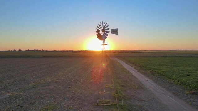 Sunset landscape, cinematic aerial tracking shot capturing old fashioned windpump spinning in the middle of arable land in La Pampa, Patagonia, Argentina.