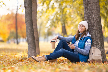 Urban golden autumn. Attractive smiling woman in warm wear sits under a tree with coffee and mobile...