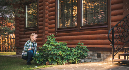 A beautiful blonde woman in a plaid shirt gardens in the sunset. A woman in gloves takes care of bushes in the backyard. Rural life. Proximity to nature.