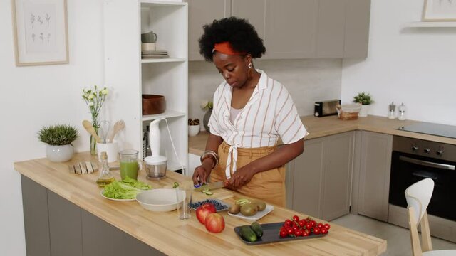 Medium slowmo shot of young African-American woman in casualwear standing by wooden kitchen table at modern apartment, cutting fresh vegetables while making healthy dinner