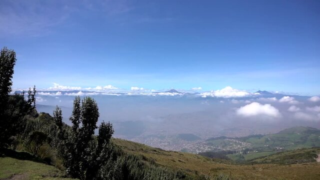 Panoramic view of the city of Quito, Ecuador, and the mountains and volcanoes that surround it. The sky is blue and there are some clouds above.