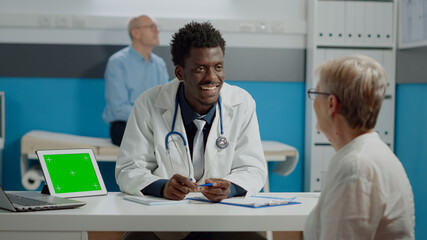 African american medic discussing with senior woman while having tablet with horizontal green screen on desk. Black doctor with chroma key template and isolated background talking to old patient