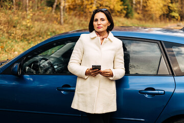 Serious fashion business woman in white female coat with  cell phone outside her car in autumn park