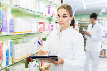 Woman specialist is attentively stocktaking medicines with notebook near shelves in pharmacy