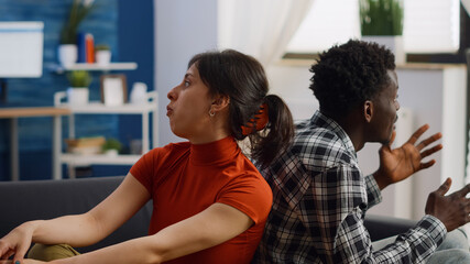 Irritated interracial couple getting into argument on sofa at home. Multi ethnic partners fighting, having relationship problems in living room. Young people sitting on couch yelling