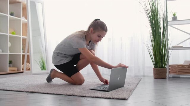 Online training. Kids sport. Video lesson. Teenager girl doing warming workout bending forward looking laptop light room interior.