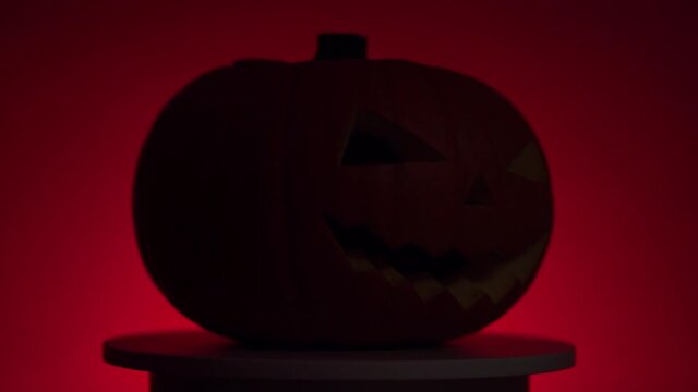 halloween orange smiling pumpkin with dynamic flashing light, carved teeth, eyes and nose on rotating circular light platform in red light on a dark background