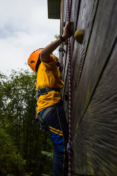A nine-year-old boy equipped with a rock-climbing gear kit is climbing a wall outdoor with confidence.
