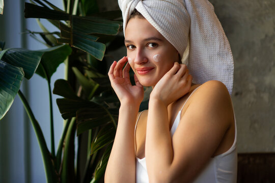 Close-up shot of a beautiful young woman doing her morning routine, applying face cream while covering her hair with a towel after washing.